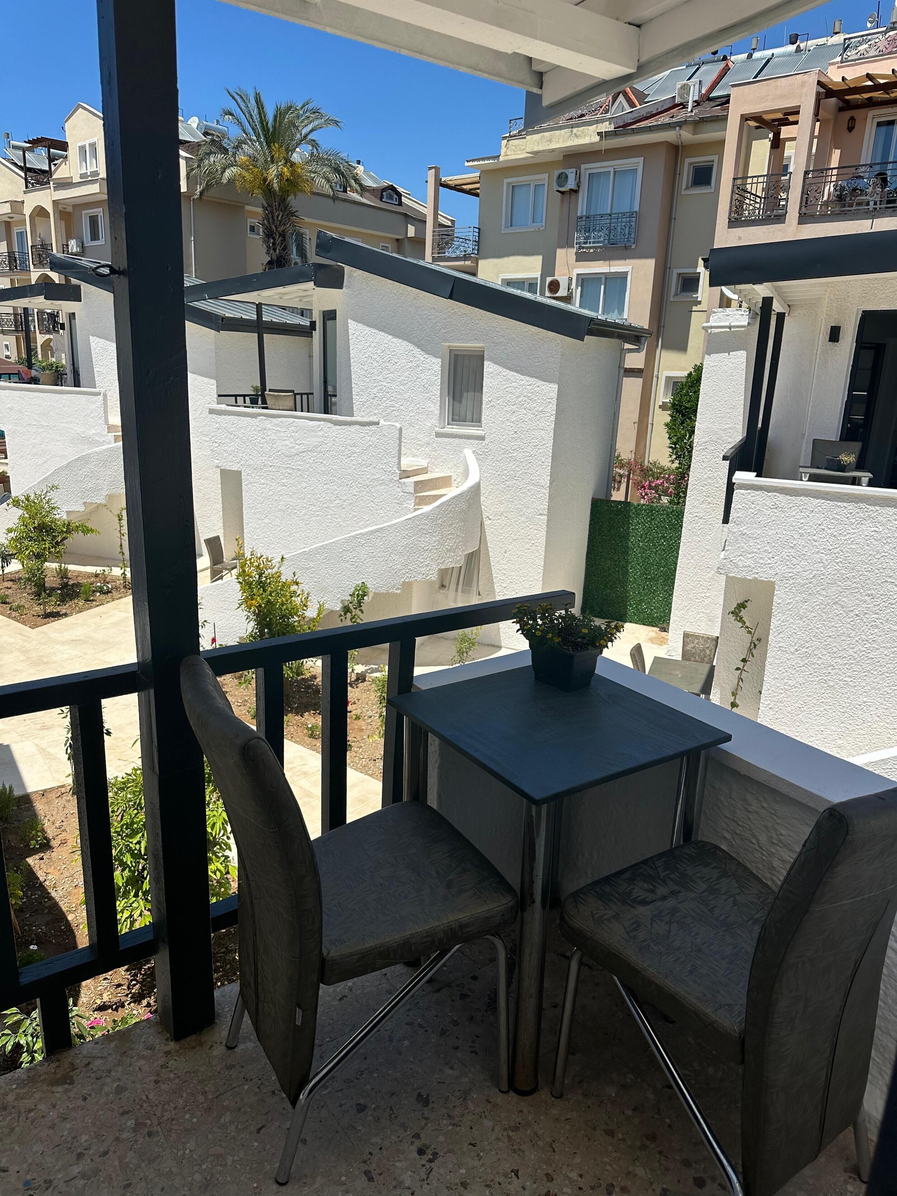 Un balcon avec une table et des chaises, donnant sur un quartier résidentiel avec des maisons et un palmier.