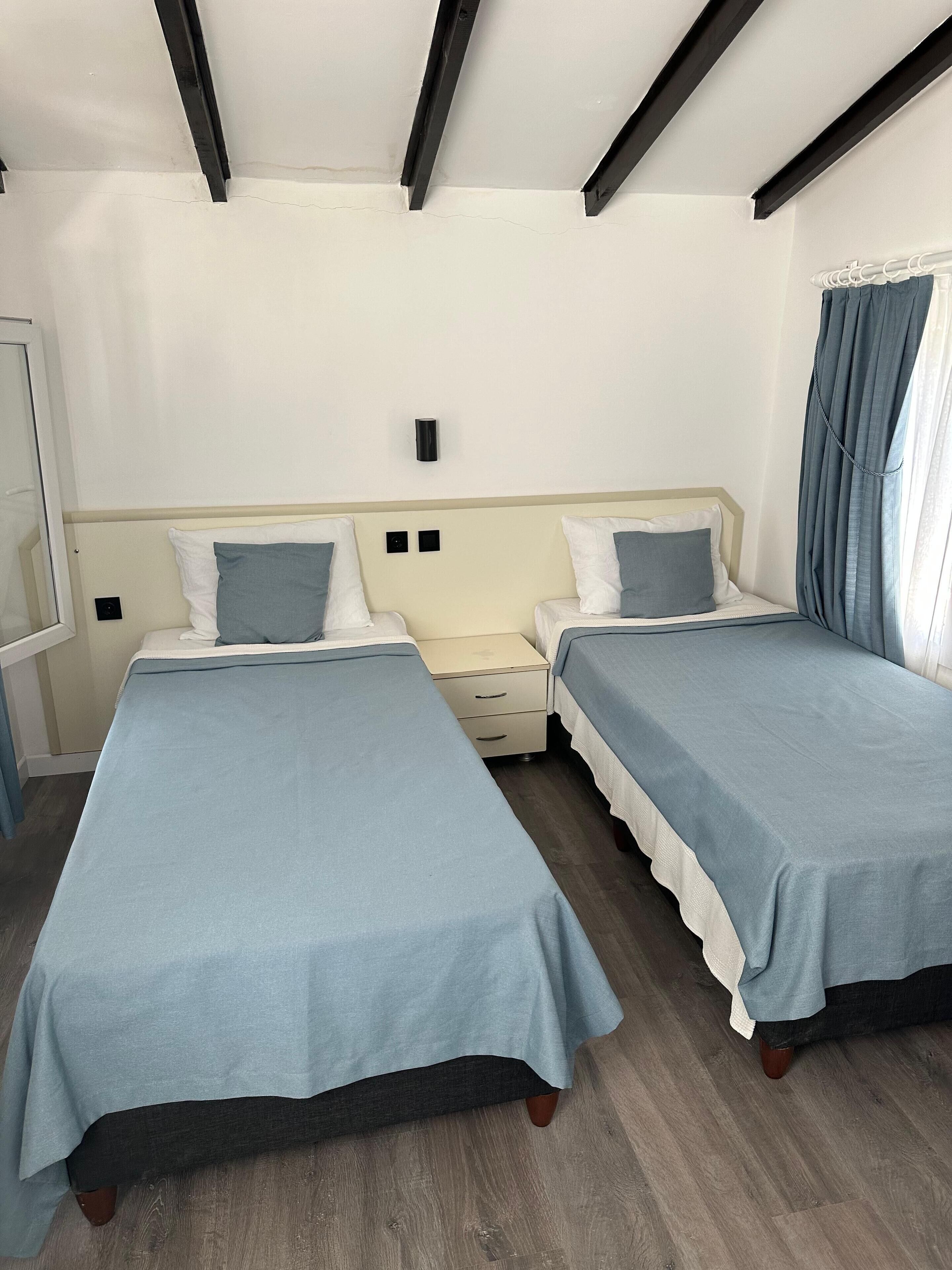 A hotel room with two single beds, wooden flooring, a white ceiling with black beams, a small bedside table, and a window with curtains.