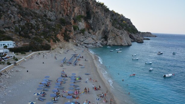 Una platja amb gandules i ombrel·les, penya-segades rocoses i embarcacions a l'aigua.