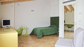 A small, well-lit room with a bed, a desk, a chair, and a television.