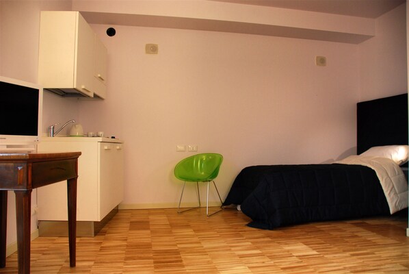 A small, sparsely furnished room with a bed, a chair, a table, and a kitchenette. - Stile Libero (Orio al Serio)