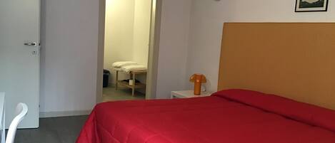 A room with a bed, a desk, and a chair. There is a door leading to another room.