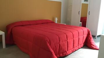 A bedroom with a large red bedspread, a white bedside table, a wardrobe, and a framed picture on the wall.