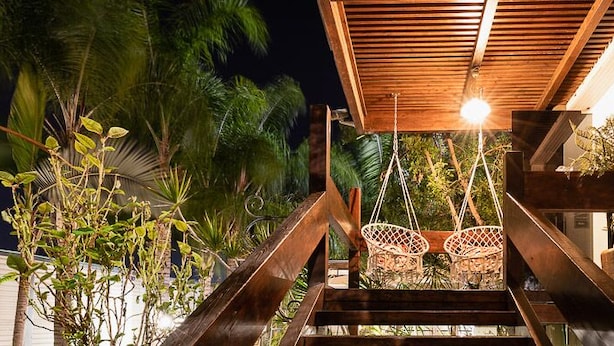 Un escalier avec deux fauteuils en osier placés sous une marquise en bois, le tout entouré de verdure.
