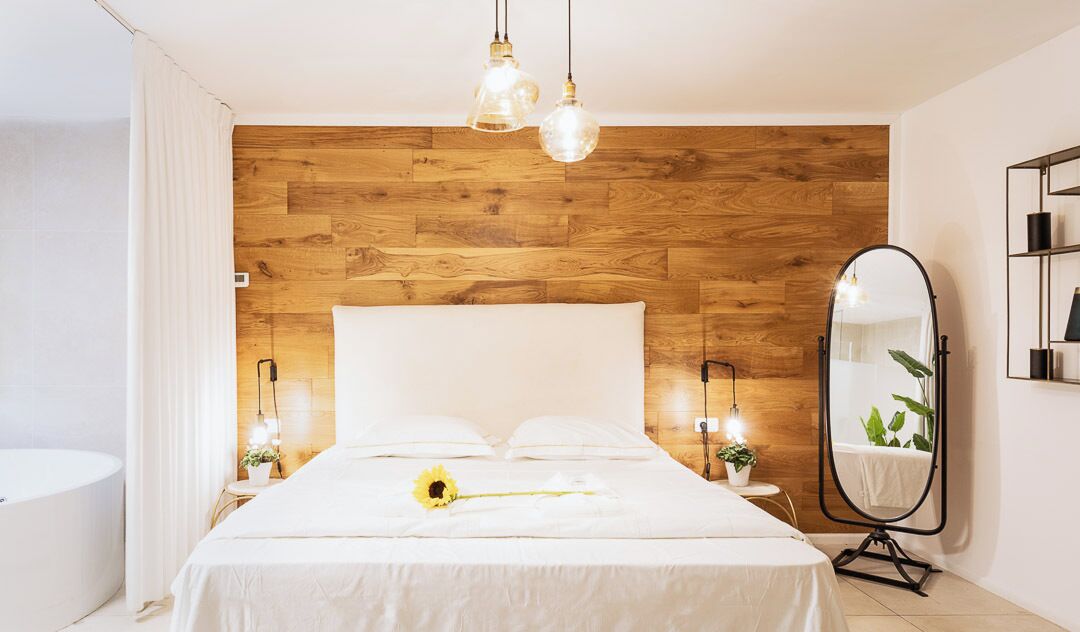 Un dormitorio moderno con cabecera de madera, una cama grande, lámparas de noche, un espejo redondo y una pequeña mesa con una planta.