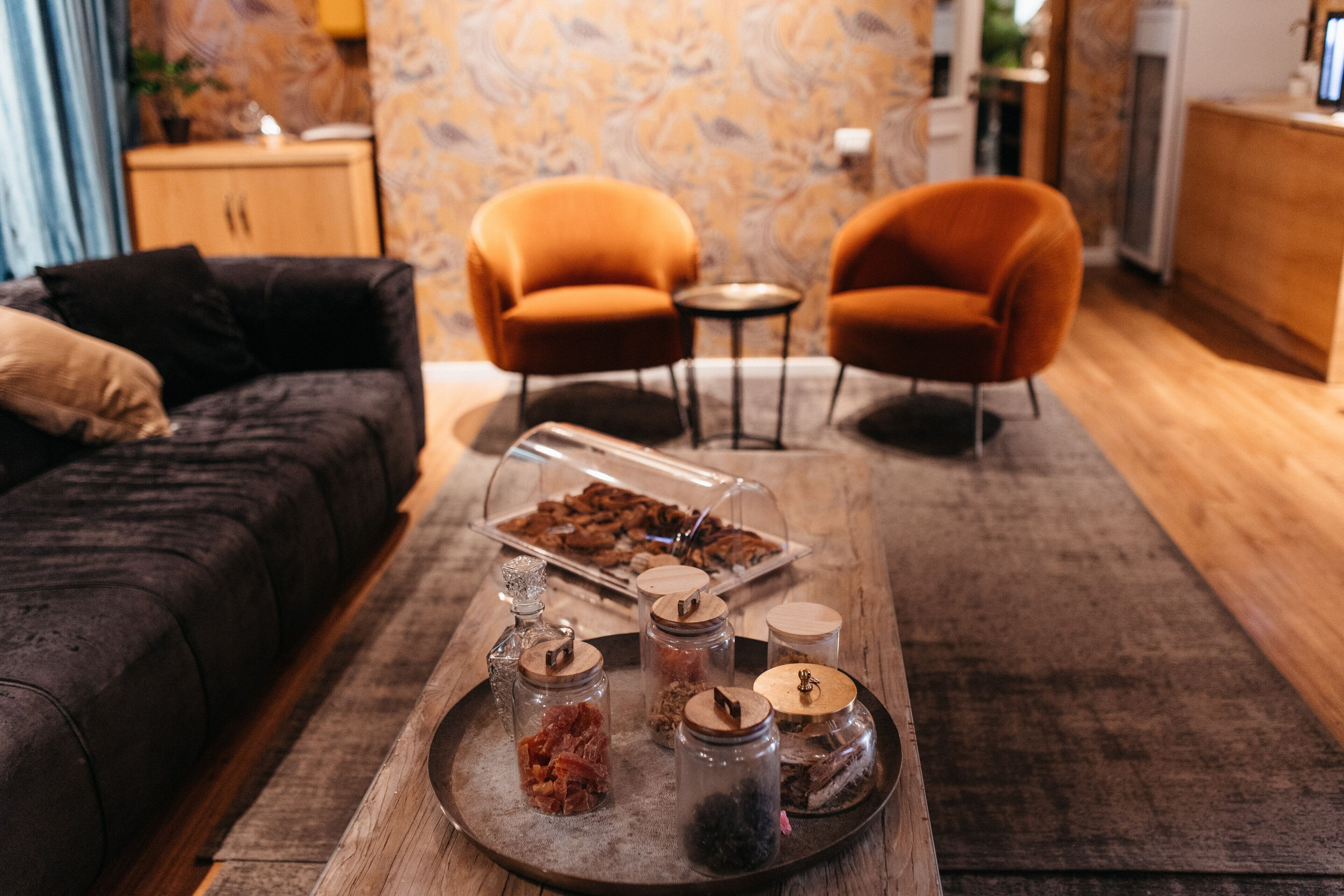 A cozy living room with a dark sofa, two orange armchairs, a wooden coffee table with jars and a tray of snacks.
