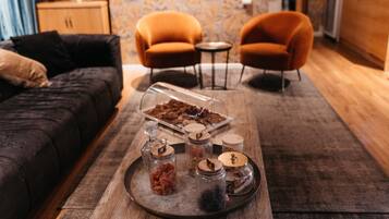 A cozy living room with a dark sofa, two orange armchairs, a wooden coffee table with jars and a tray of snacks.