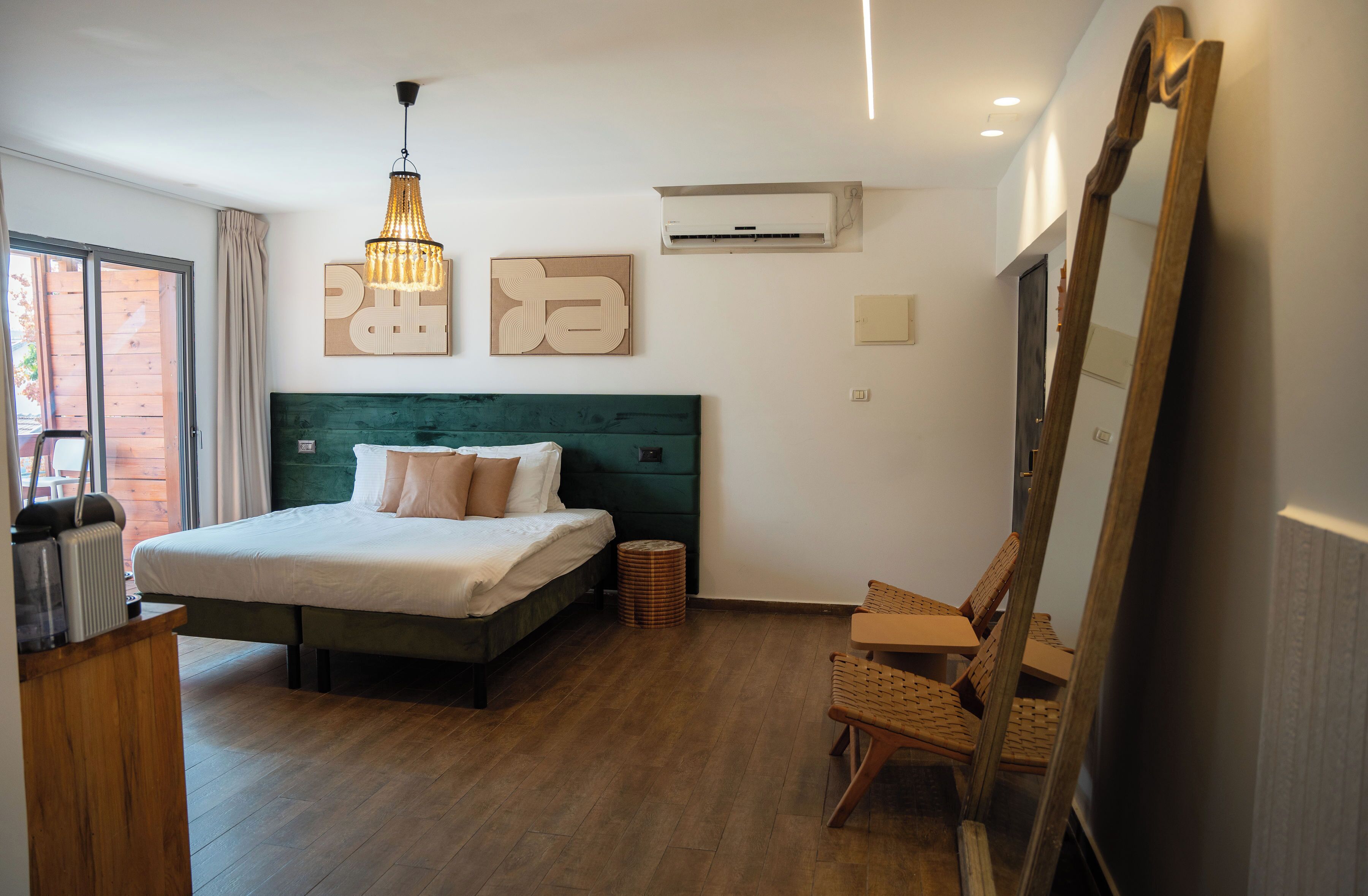 A bedroom with a bed, a wooden floor, a hanging light fixture, a wall-mounted air conditioner, and a large mirror.
