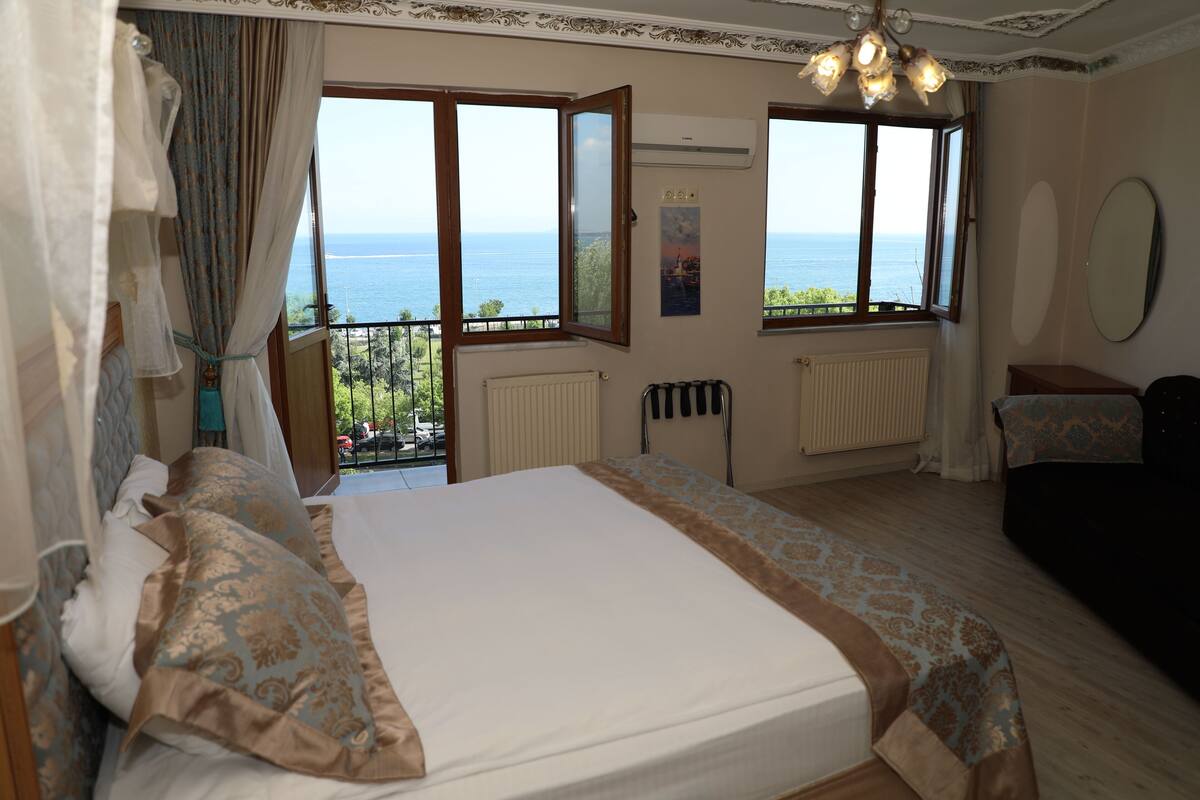 A bedroom with a large bed, two windows with ocean views, a radiator, and a ceiling light fixture.