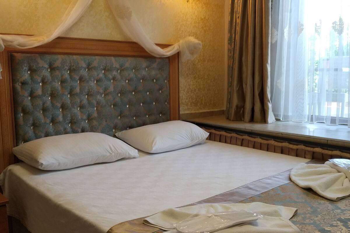 A hotel room with a tufted headboard, a large bed with white linens, a window with sheer curtains, and a bedside table with towels.