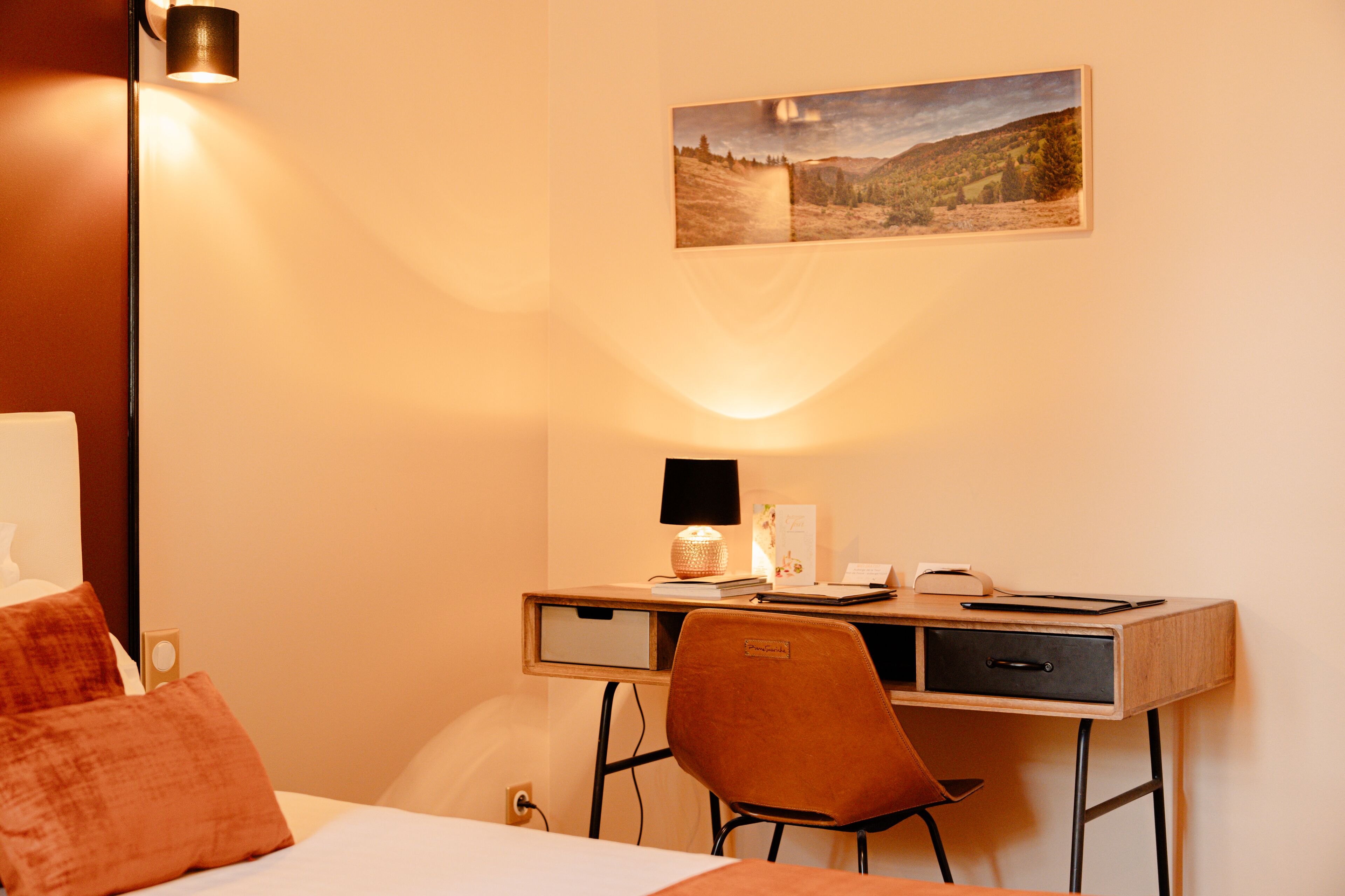 Een hotelkamer met een bed, een bureau met een lamp, een stoel en een ingelijst kunstwerk aan de muur.