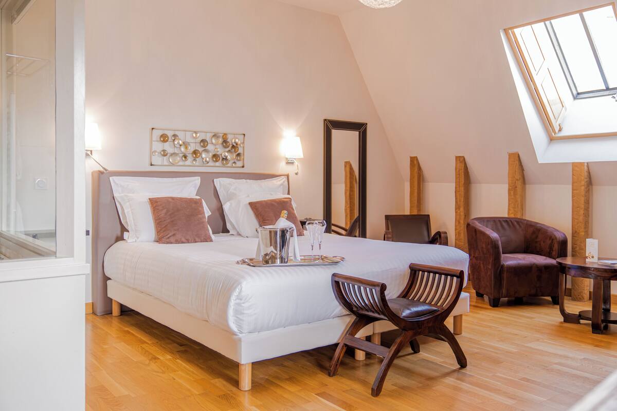 A hotel room with a large bed, a wooden chair, a small table, and a skylight.