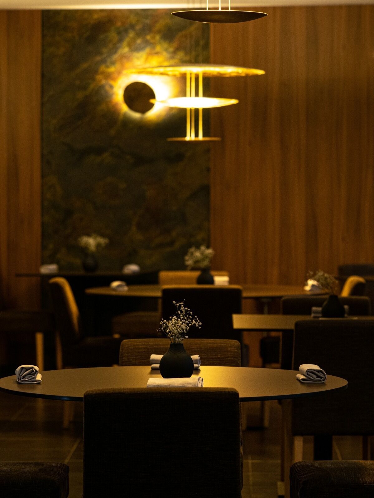 Un restaurante tenuemente iluminado con mesas y sillas redondas, un jarrón con flores en cada mesa y una pared con una lámpara circular.