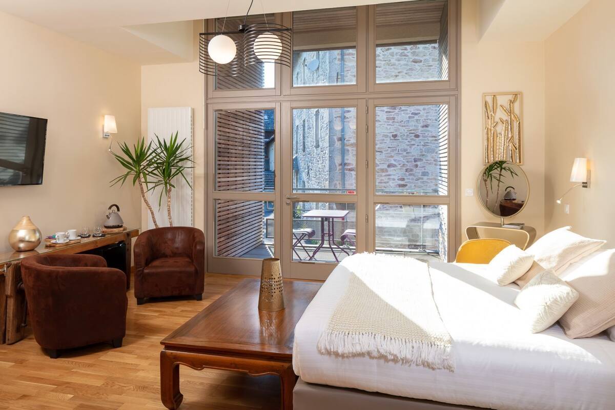 A modern hotel room with a large window, a wooden desk, a sofa, and a dining table.