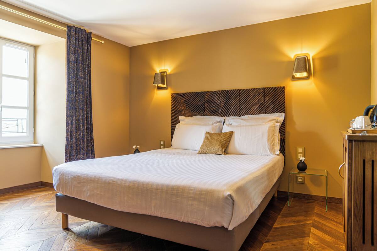 A neatly made bed with a headboard, two wall sconces, a small table with a vase, and a wooden cabinet.