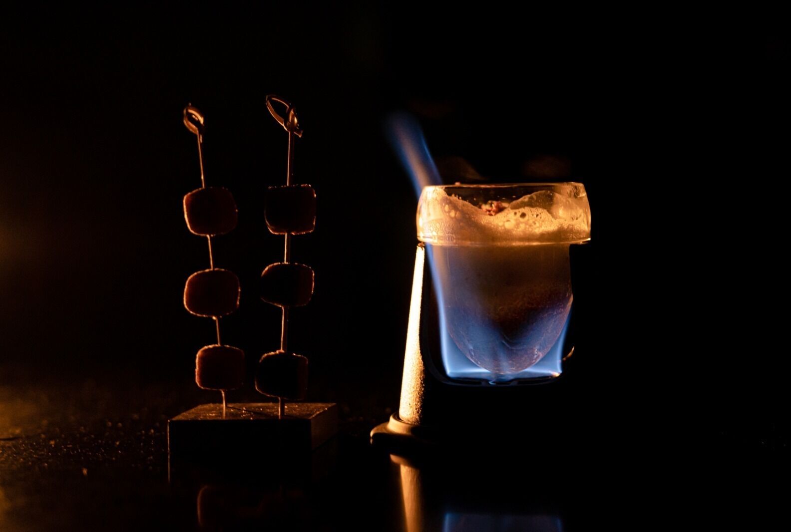 Een glas met een donkere drank en een blauwe vlam, mogelijk een warme drank of een cocktail die wordt bereid.