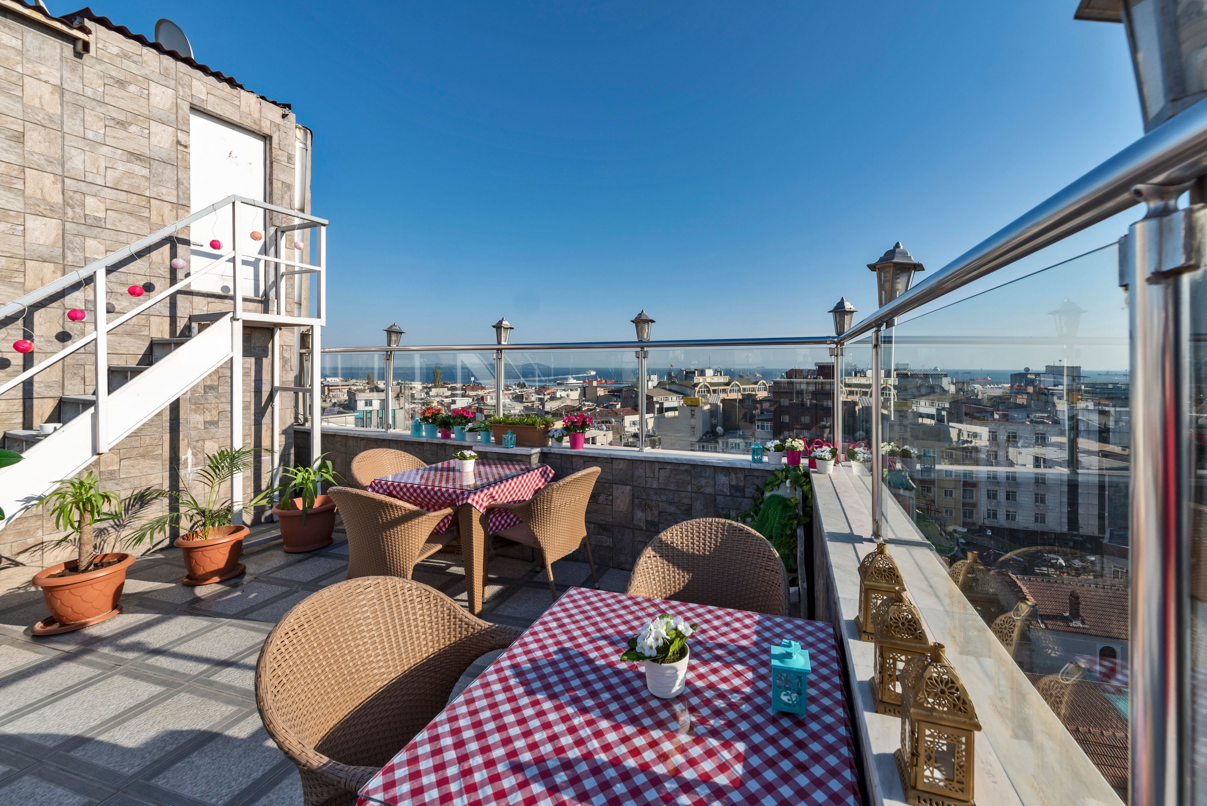 A rooftop terrace with wicker furniture, a checkered tablecloth, potted plants, and a city view.