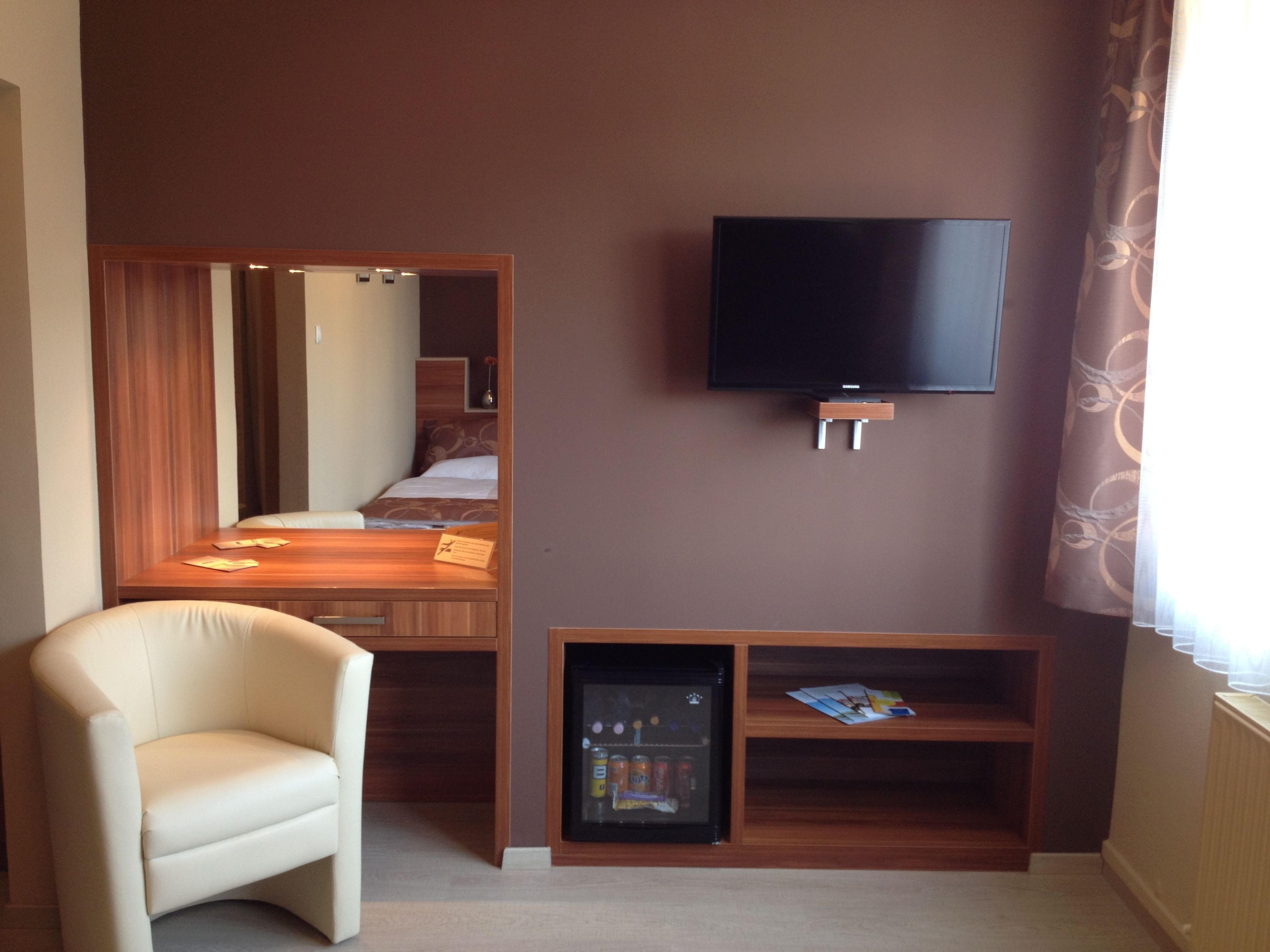 A hotel room with a bed, a TV mounted on the wall, a small refrigerator, and a seating area with a chair.