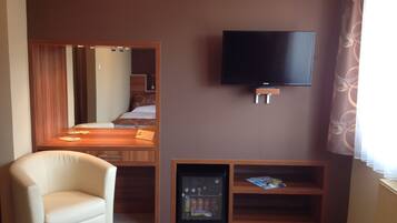 A hotel room with a bed, a TV mounted on the wall, a small refrigerator, and a seating area with a chair.