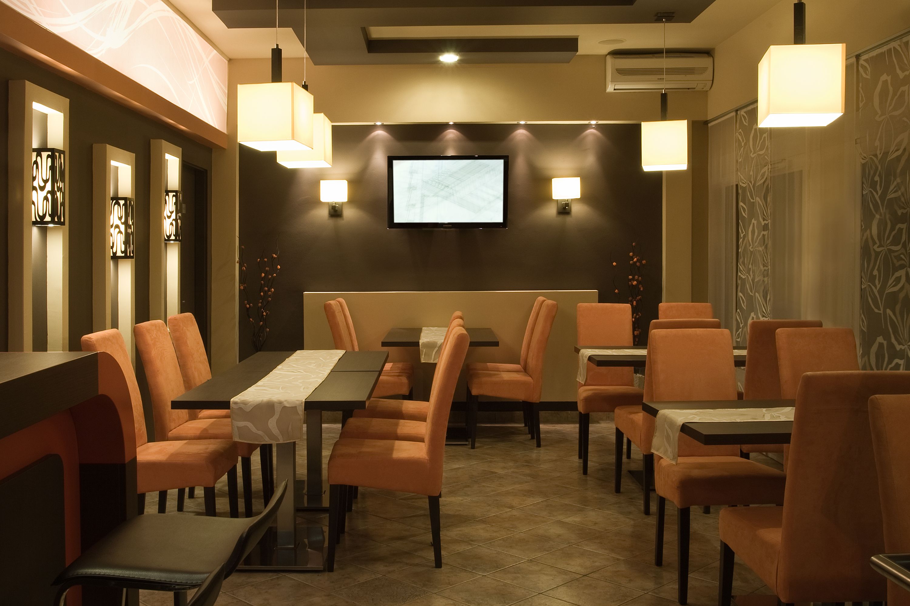 A restaurant with a long table and orange chairs, a TV screen on the wall, and pendant lights.