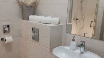 A modern bathroom with a white sink, toilet, and shower. There is a towel rack with towels and a decorative vase on the wall.