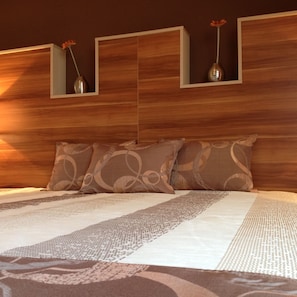 A neatly made bed with patterned bedding, a wooden headboard, and two vases with flowers on the wall above. - Hotel Alfa (Gyor)
