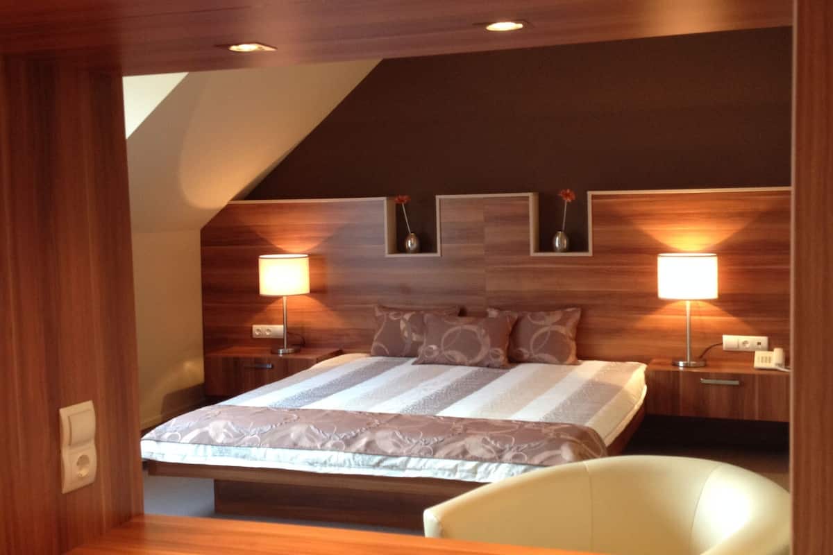 Une chambre d’hôtel moderne avec un plafond en bois, un grand lit recouvert d’une courtepointe à motifs, deux lampes de chevet et une chaise blanche.