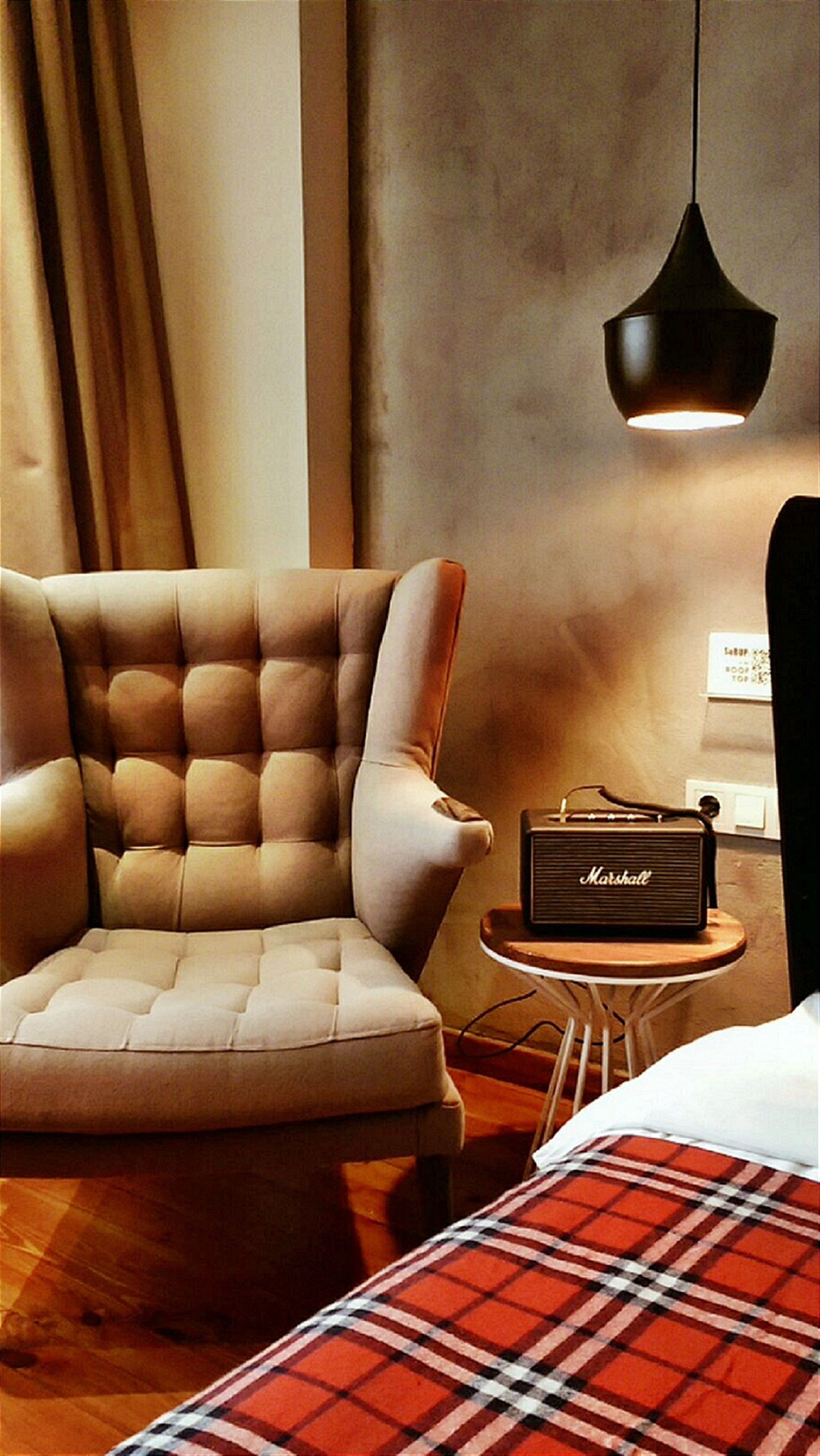 A modern hotel room with a tufted armchair, a small side table with a vintage radio, and a plaid blanket on the bed.
