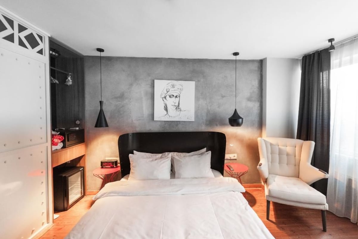 A modern hotel room with a large bed, a side table, a chair, a painting on the wall, and a wardrobe.