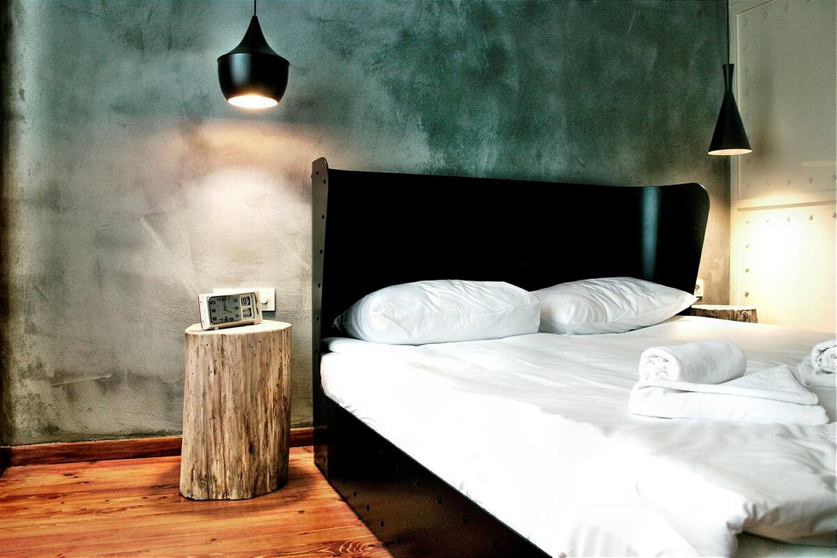 Un dormitorio con suelo de madera, una cama con ropa de cama blanca, una mesita de noche con un reloj y dos lámparas colgantes.
