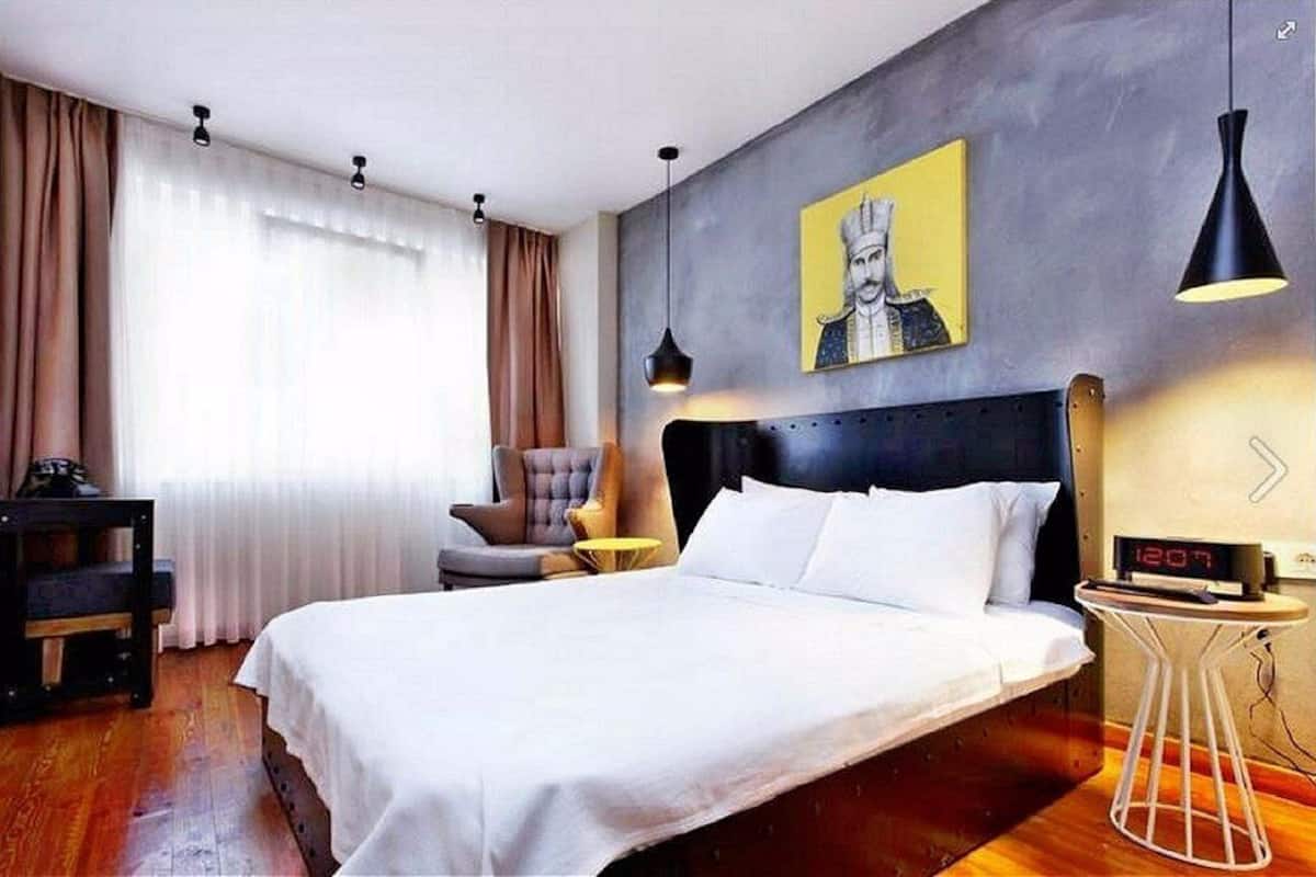 Habitación de hotel con una cama grande, una silla, una mesita y un reloj de pared, además de un cuadro en la pared.