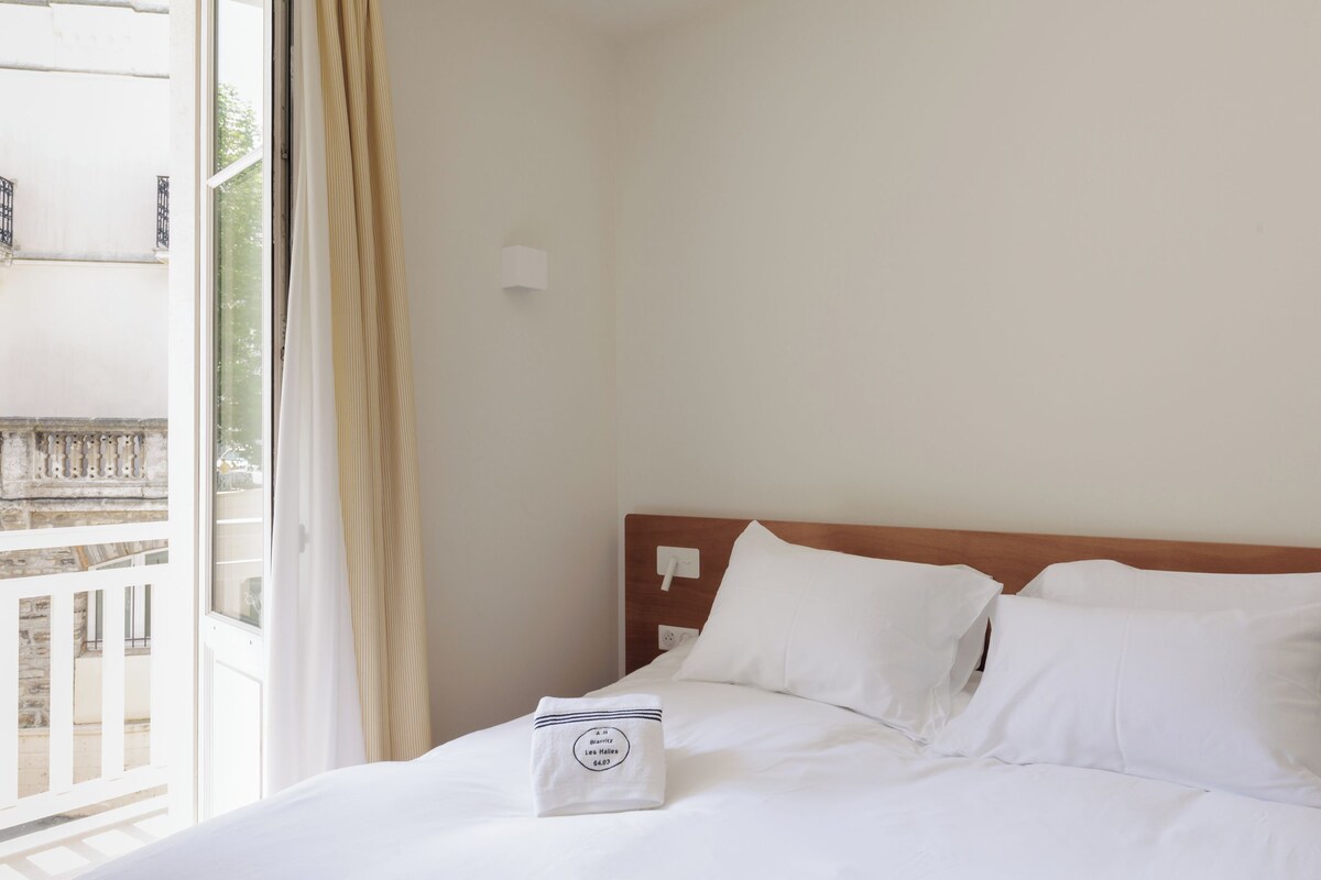 A hotel room with a bed, white linens, a window with curtains, and a view of a balcony.