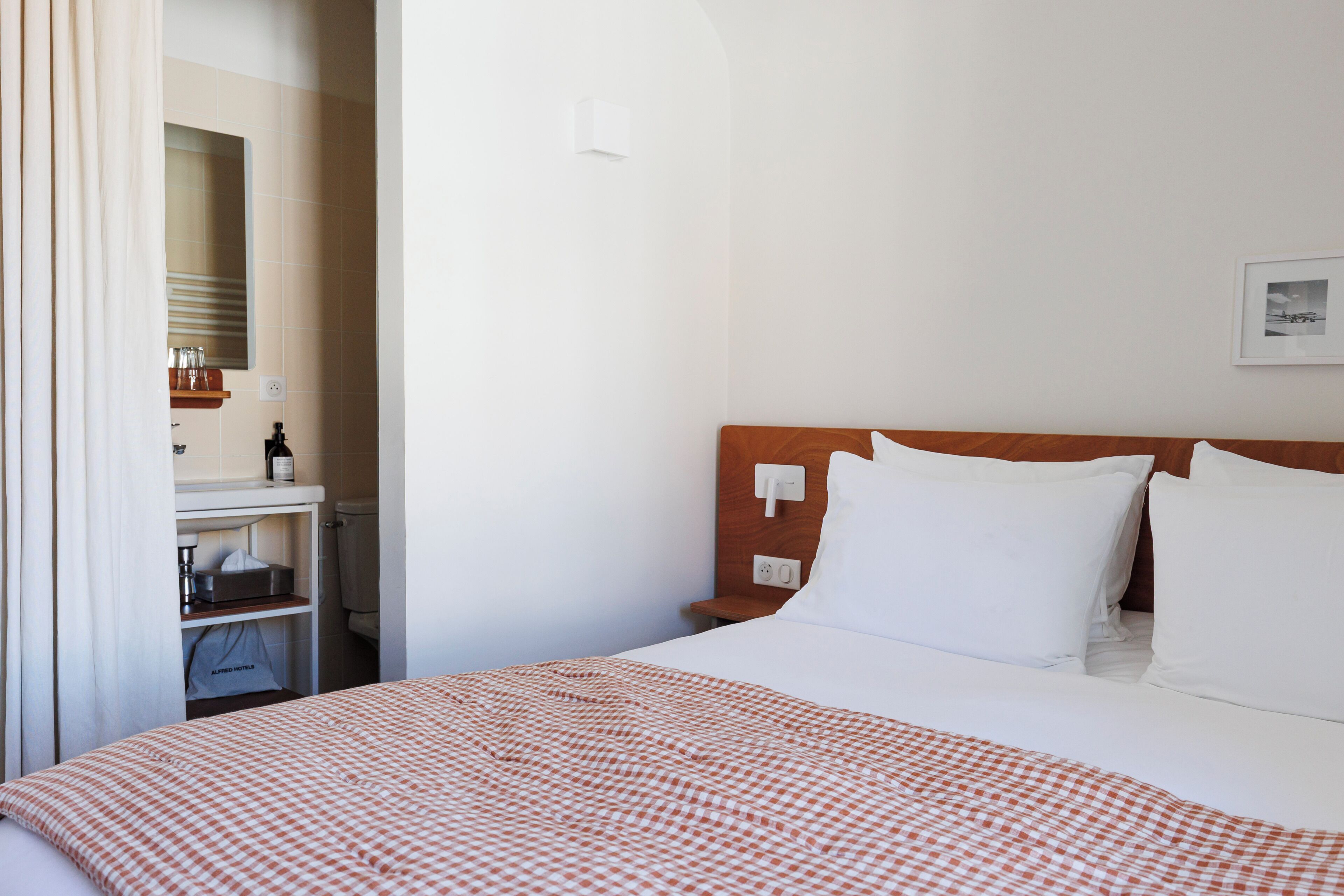 Une chambre d’hôtel avec un lit, une table de chevet, un miroir, une étagère avec des articles de toilette et un tableau encadré au mur.