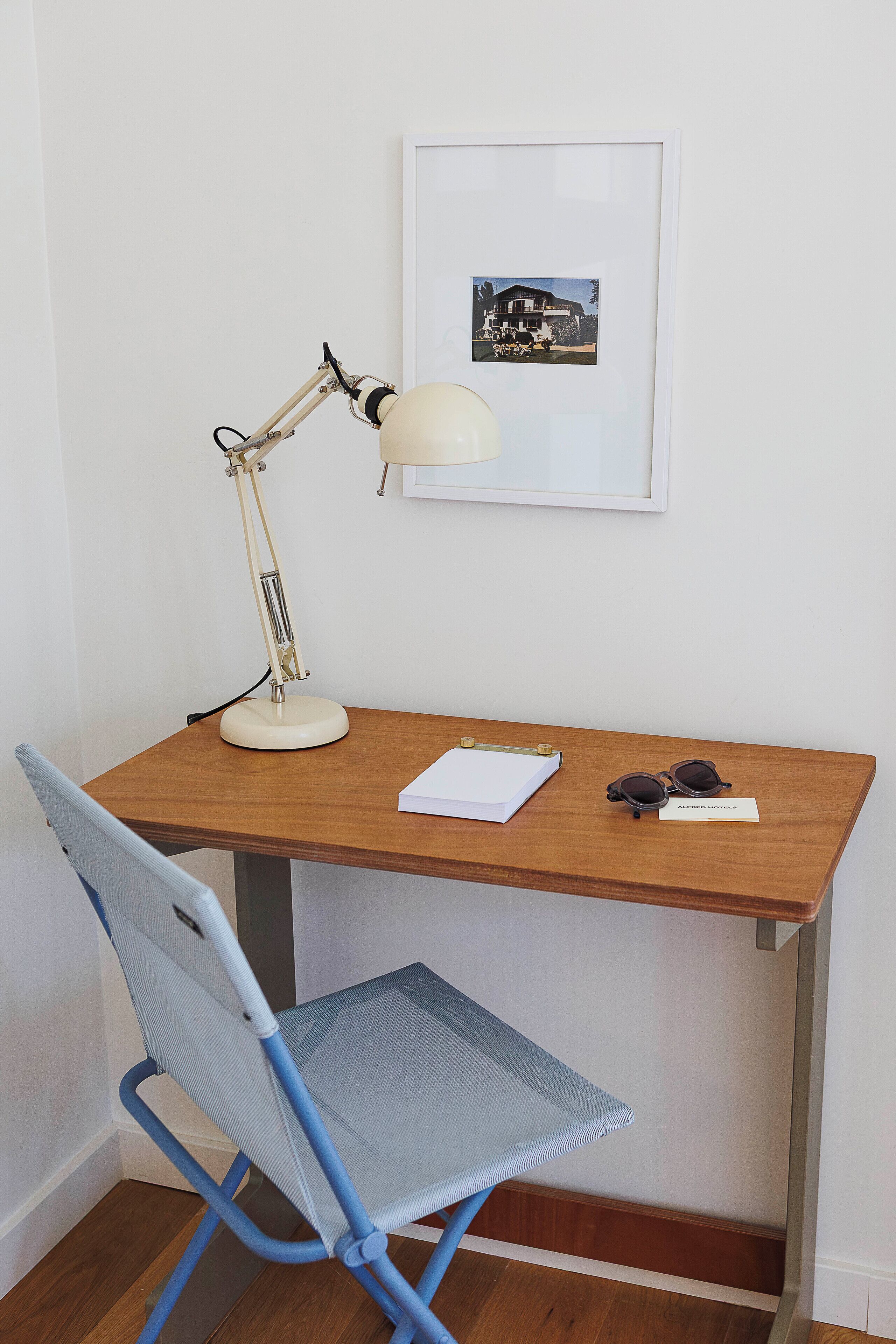 Un bureau en bois avec une lampe de bureau blanche, une chaise bleue, un tableau encadré accroché au mur et un carnet sur lequel reposent des lunettes de soleil.