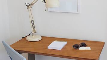Un bureau en bois avec une lampe de bureau blanche, une chaise bleue, un tableau encadré accroché au mur et un carnet sur lequel reposent des lunettes de soleil.