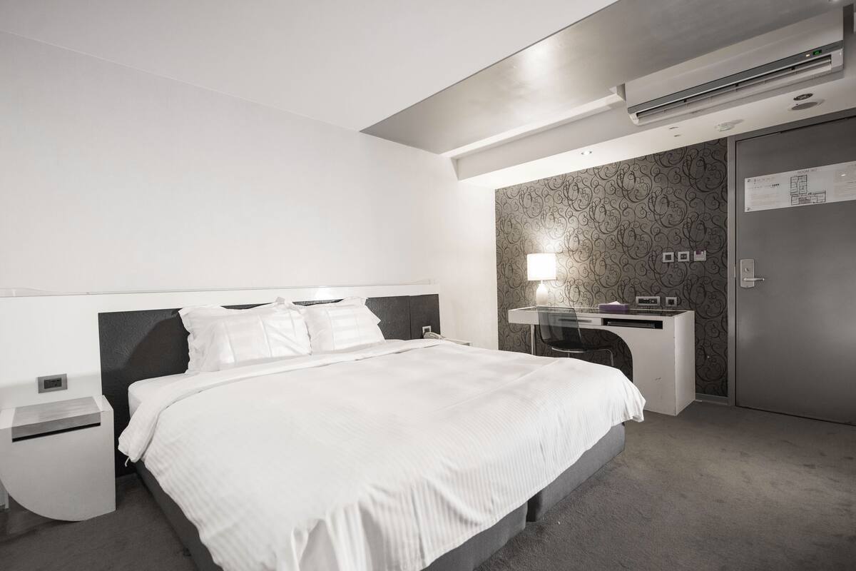 A modern hotel room with a large bed, a desk with a lamp, and a wardrobe.