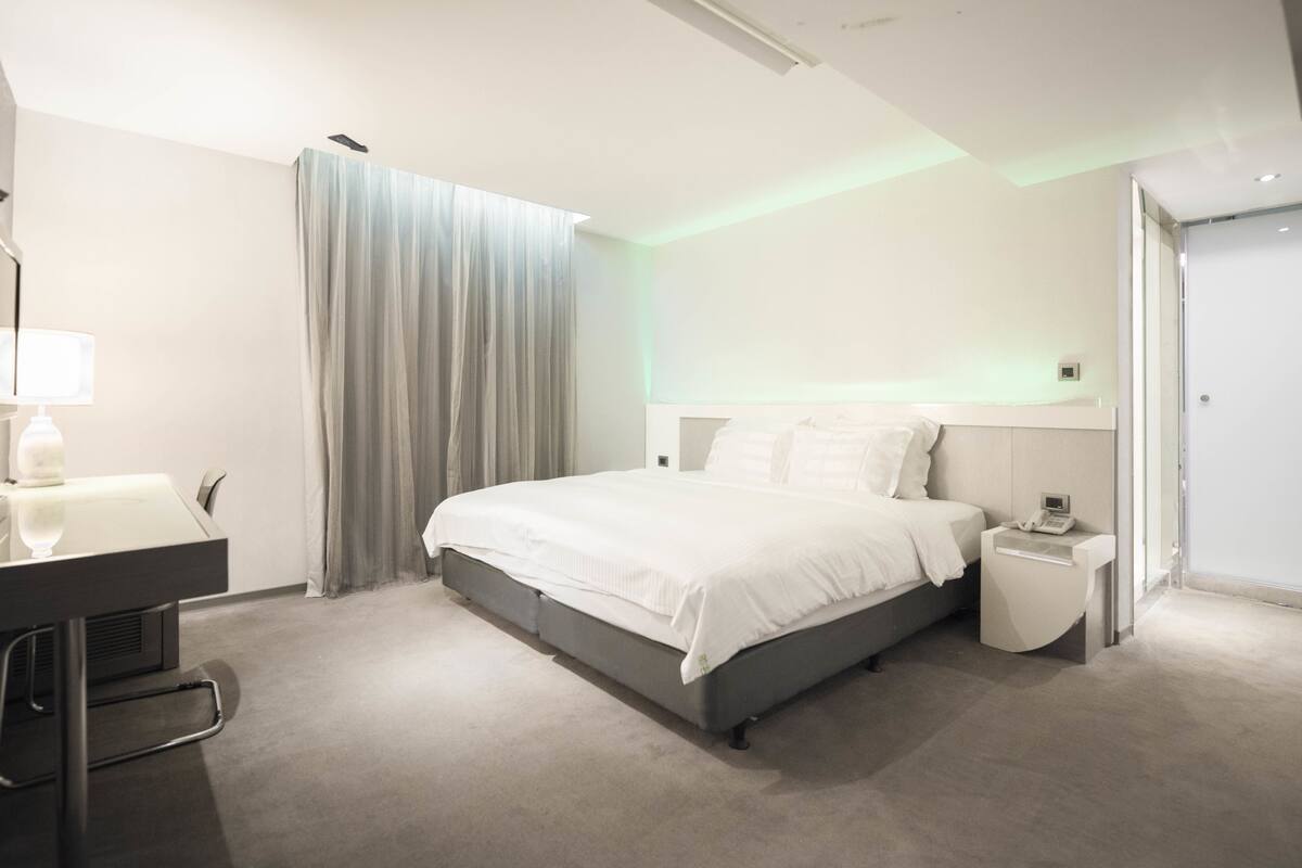 A modern hotel room with a large bed, bedside table, and a desk with a chair.