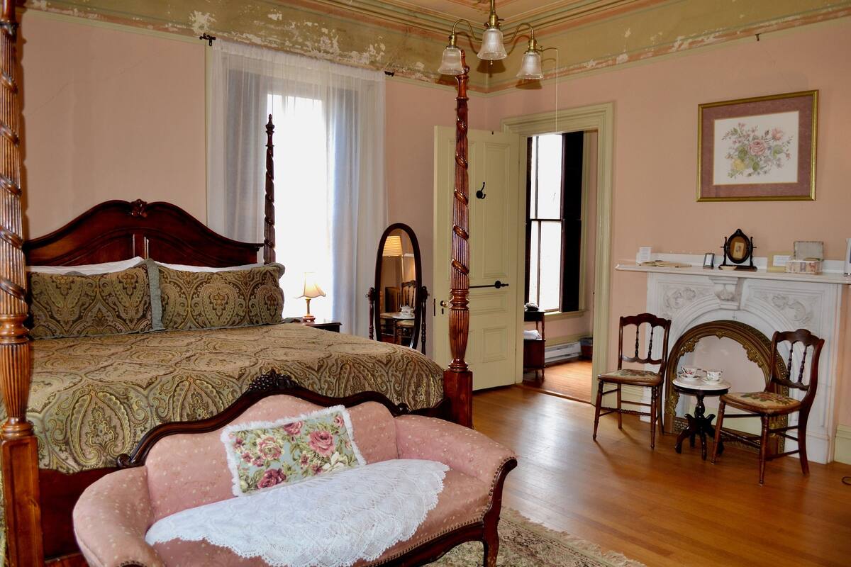 A bedroom with a four-poster bed, a chaise lounge, two chairs, a fireplace, and a mirror.