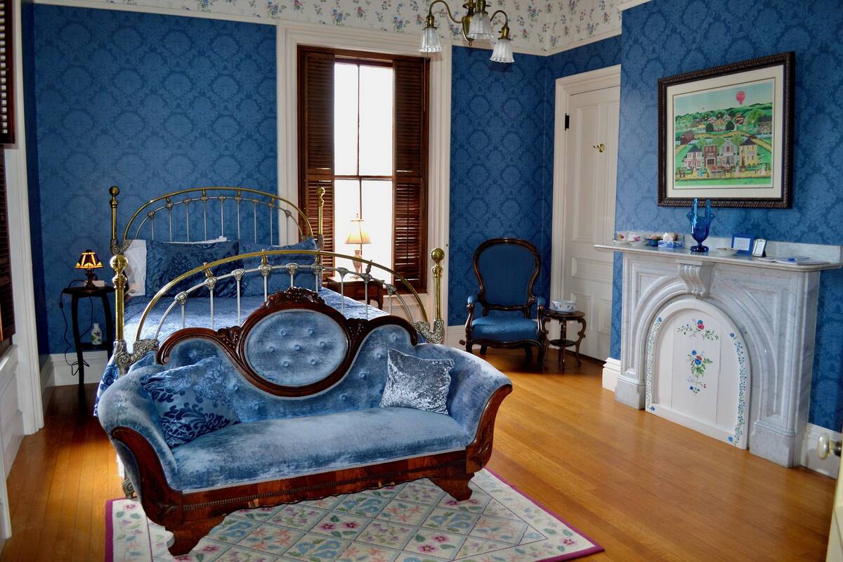 A bedroom with a bed, a blue sofa, a fireplace, and a painting on the wall.