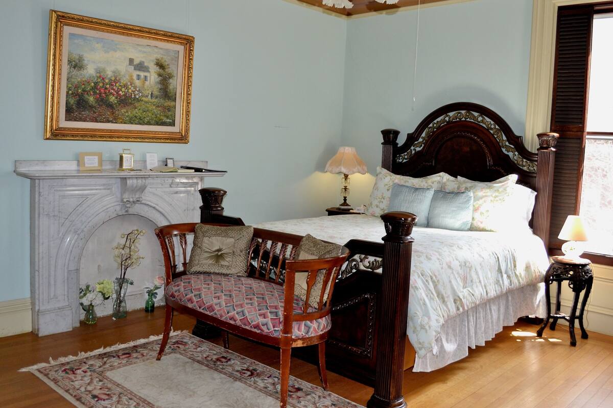 A bedroom with a bed, a chair, a fireplace, and a painting on the wall.