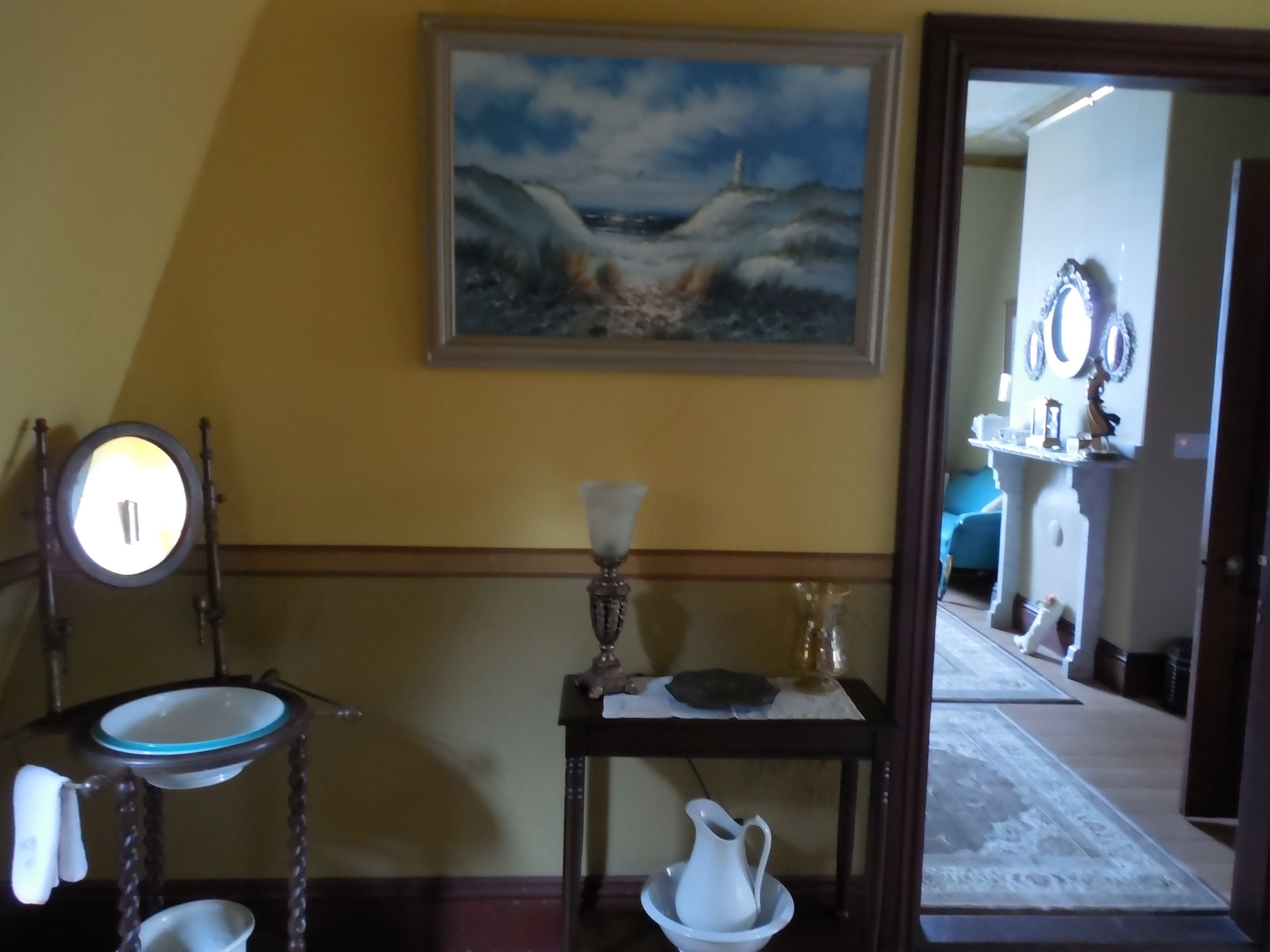 A room with a painting of a seascape, a mirror, a sink, a small table with a pitcher, and a rug.