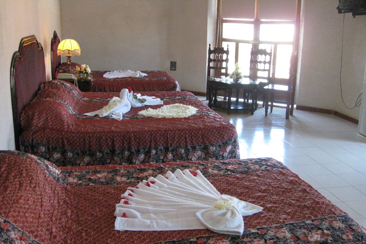 Ein Hotelzimmer mit zwei Betten, jedes mit floralem Überwurf und weißen Handtüchern, die fächerförmig drapiert sind. Ein Holzstuhl und ein kleiner Tisch mit Lampe und Blumen gehören zur Einrichtung.