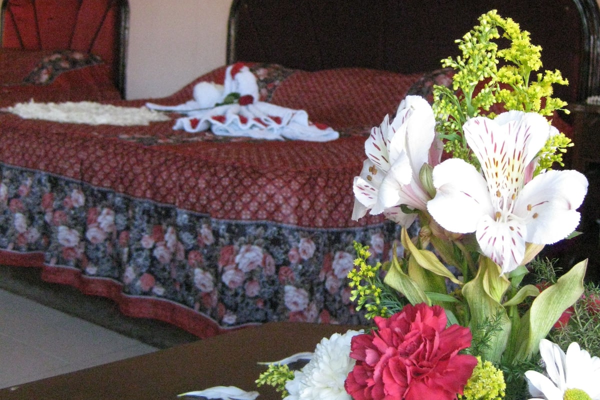 Ein Bett mit floraler Tagesdecke, ein Tisch mit Blumenarrangements und ein Sessel mit einem Blumenkissen.