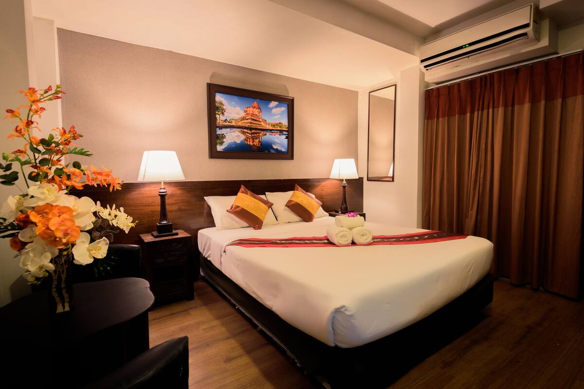 A hotel room with a bed, two bedside lamps, a nightstand, a vase of flowers, a painting on the wall, and a window with curtains.