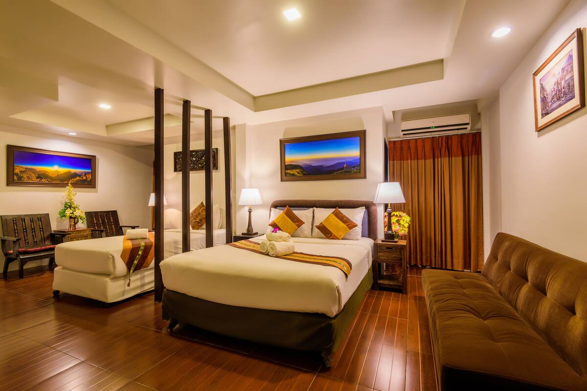 A hotel room with a large bed, a sofa, a desk, and a chair. There are paintings on the walls and an air conditioning unit.