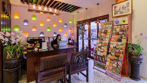 Un espace d’accueil avec un bureau en bois, une réceptionniste, un mur agrémenté de fenêtres illuminées, un présentoir de cartes postales et une plante en pot.