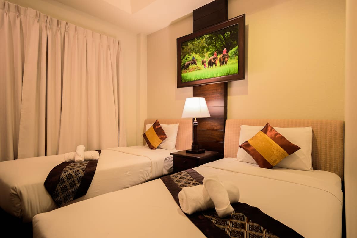A hotel room with two beds, a TV mounted on the wall, a nightstand with a lamp, and a window with curtains.