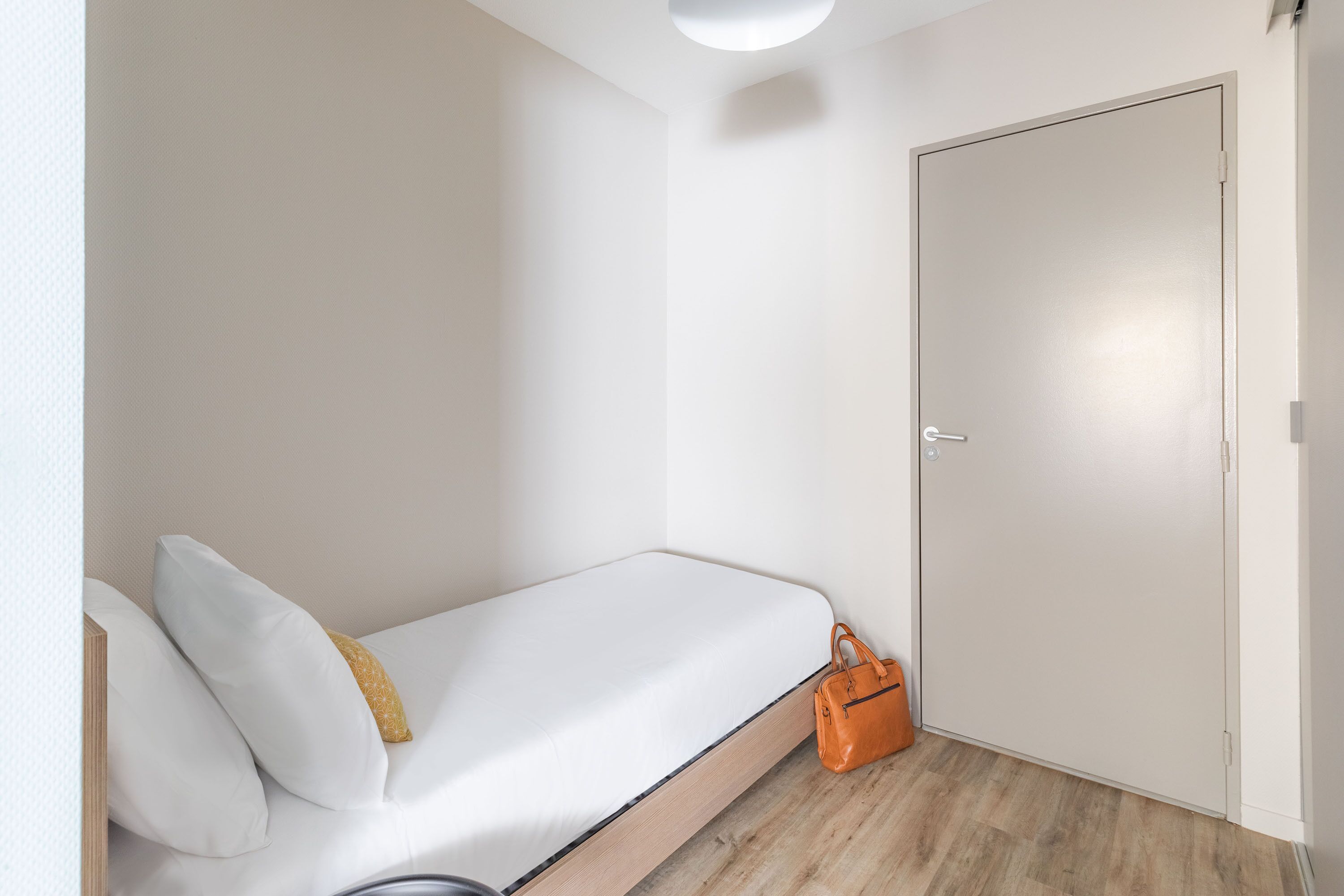 Habitación individual con una cama, piso de madera, pared blanca y puerta cerrada.