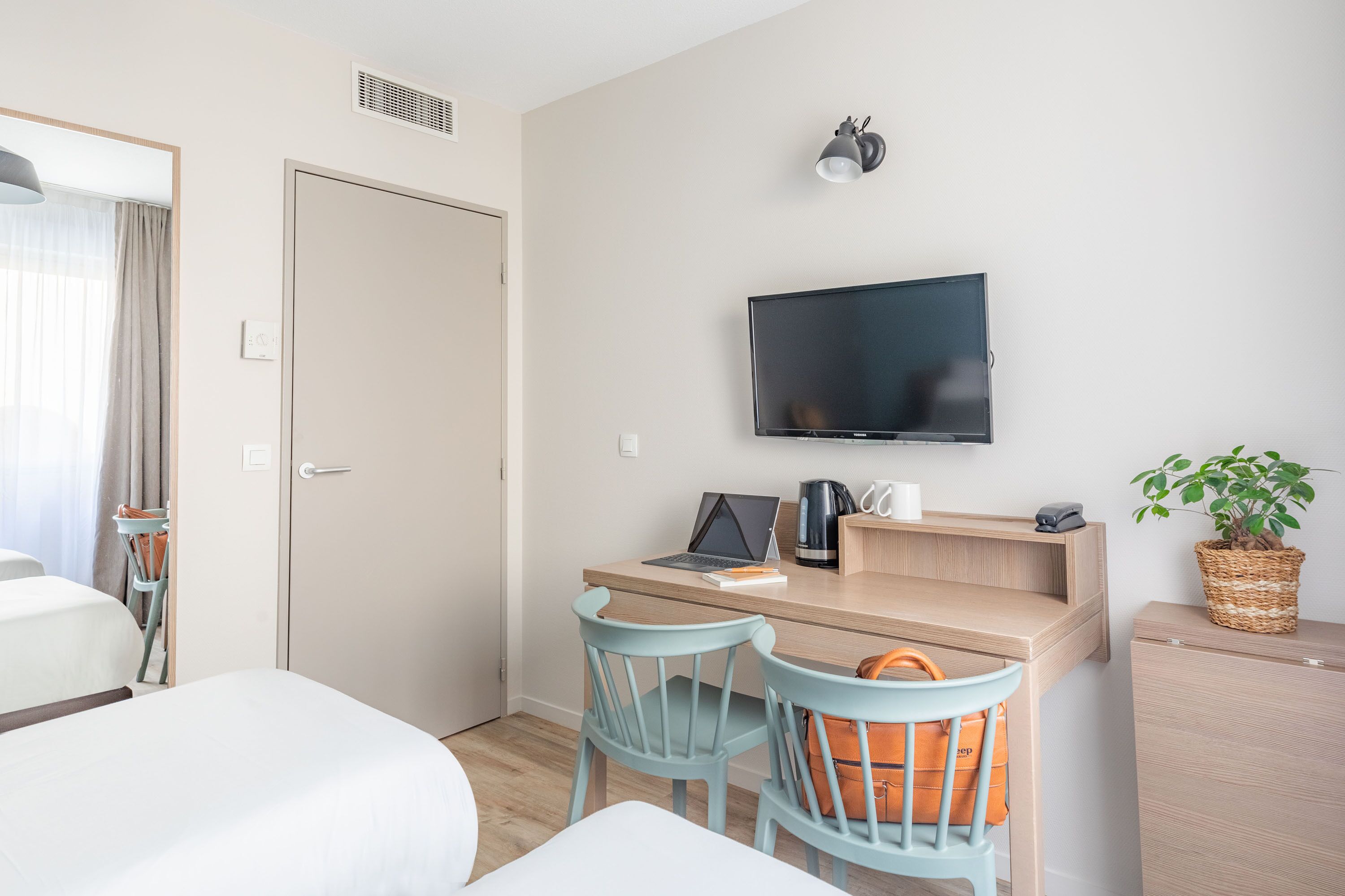 A hotel room with a bed, a desk with a laptop, a TV, and a plant.