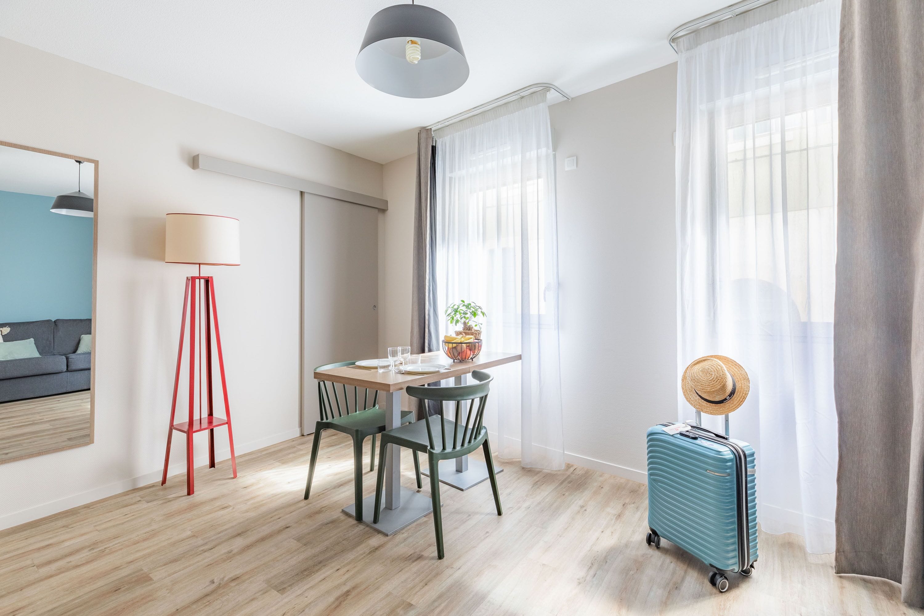 Quarto de hotel moderno com área de refeições, luminária de pé, planta e mala.